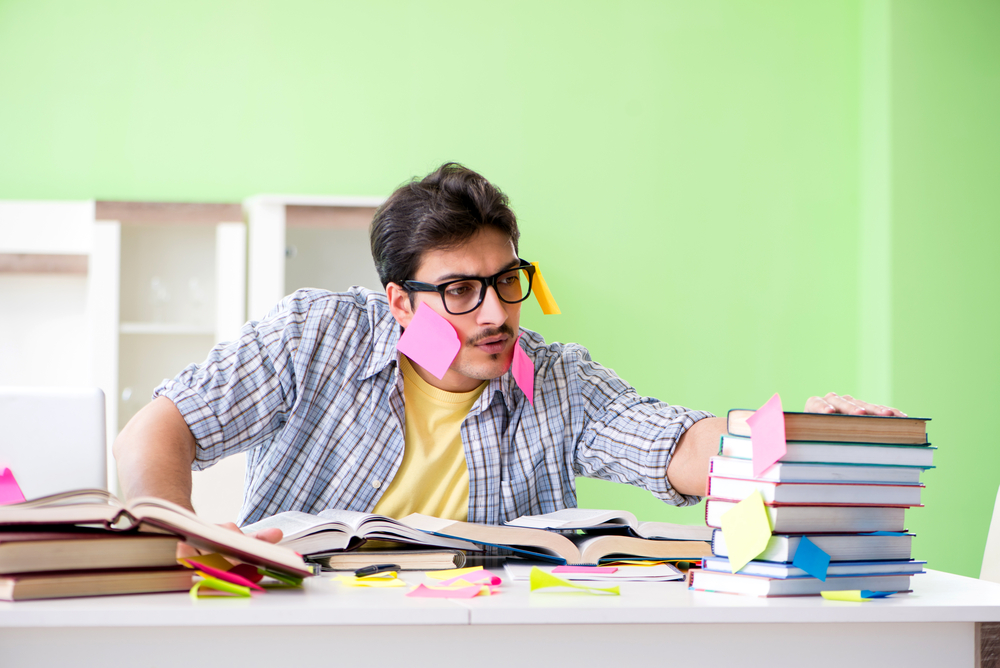 A harried man in a blue plaid shirt over a yellow tshirt and covered in post-it notes reaches for a pile of books on a cluttered desk. He is in a room with lime green walls and empty white bookshelves